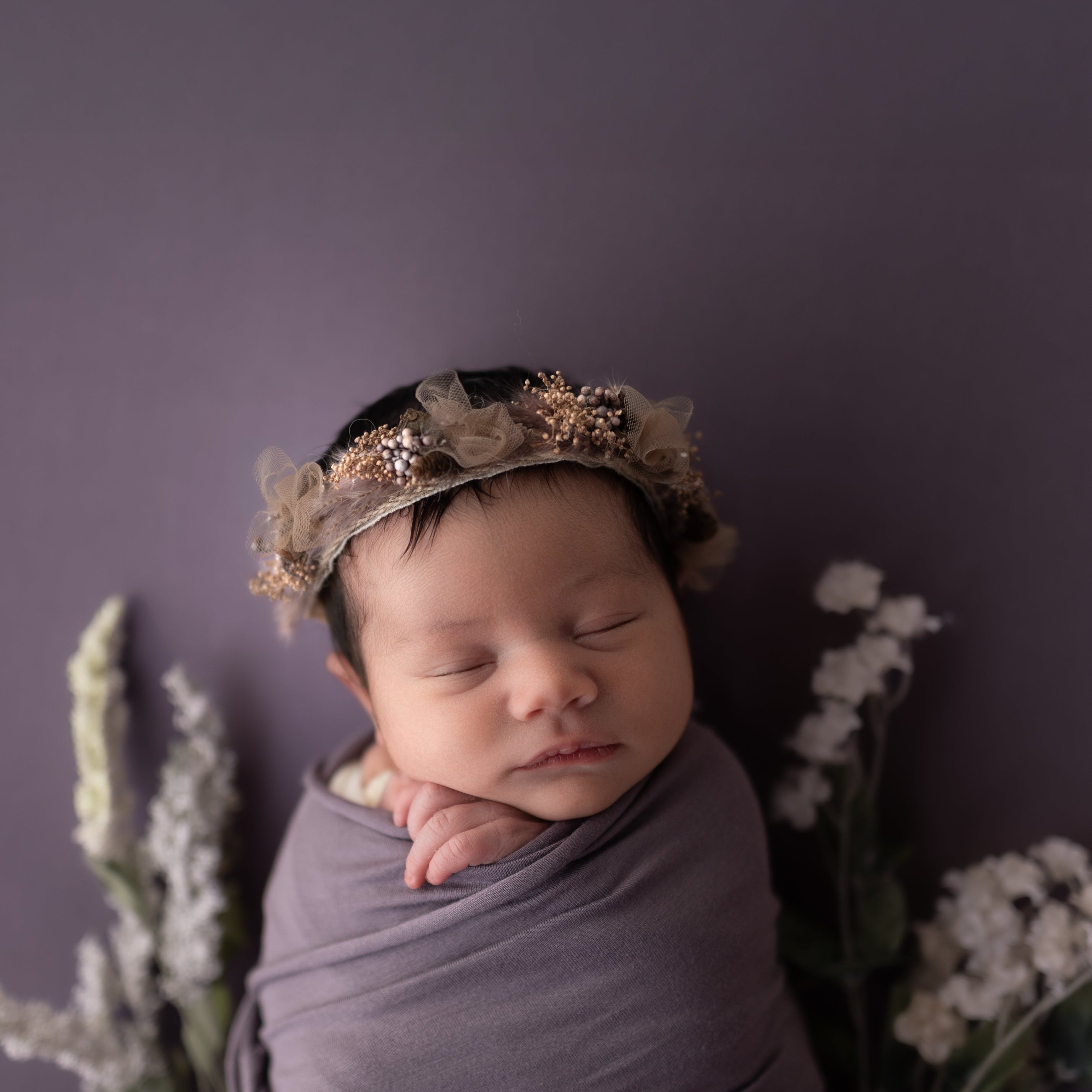 Newborn baby wrapped in purple fabric with a floral headband against a purple background