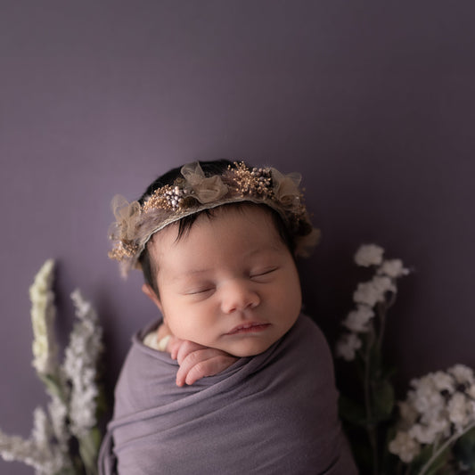 Newborn baby wrapped in purple fabric with a floral headband against a purple background
